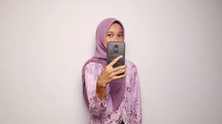 Photo for Indonesian teenage girls wearing kebaya and hijab pose holding smartphone selfies on an isolated white background - Royalty Free Image