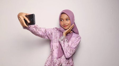 Indonesian teenage girls wearing kebaya and hijab pose holding smartphone selfies on an isolated white background