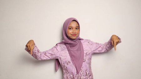 Lächelnde indonesische Teenagermädchen in Kebaya und Hijab posieren nach unten zeigend auf einem isolierten weißen Hintergrund