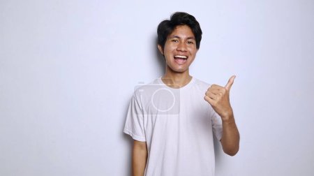 excited young asian man in white shirt with funny expression showing thumbs up okay, great, cool, steady, winning isolated white background