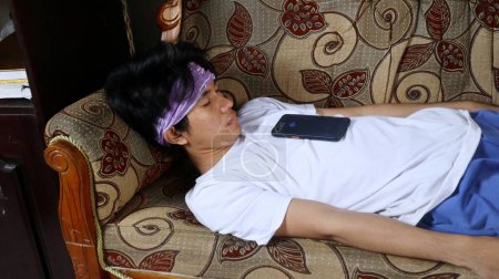 joven asiático hombre durmiendo en sofá con smartphone en su pecho