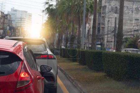 Photo for Traffic jam on the road in the city at sunset. Close up rear side of red car with turn on brake light. Beside with island of green grass and trees. And city for background. - Royalty Free Image