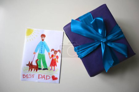 Photo for Photo of postcard for father drawn by child. Gift of blue color with bow. White background. Drawing with pencils. Dad is depicted with girl and dog. Girl eating ice cream. Inscription to the Best Dad. - Royalty Free Image