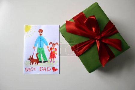 Photo for Photo of postcard for father drawn by child. Gift of green color with red bow. White background. Drawing with pencils. Dad depicted with girl and dog. Girl eating ice cream. Inscription to Best Dad. - Royalty Free Image