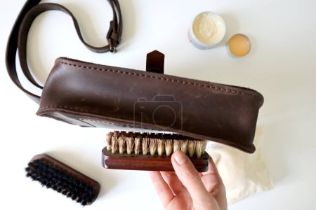 Photo of the process of cleaning a leather bag. A human hand holds a brush. Cleans the bottom of the bag. Crazy Horse brown leather. Has vintage scratches. Brush, wax, sponge around. White background.
