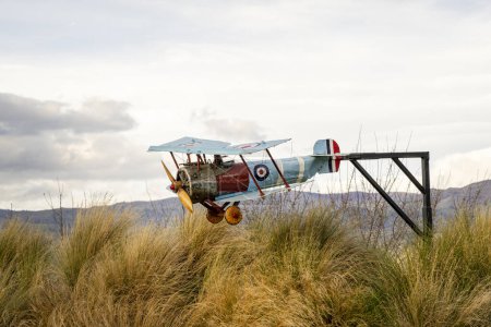 Photo for Cromwell, New Zealand - May, 2023. Enchanting scene of vintage model airplane in blue, yellow, and red hues. Flying above tall grass, framed by majestic mountains and a clear sky with scattered clouds - Royalty Free Image