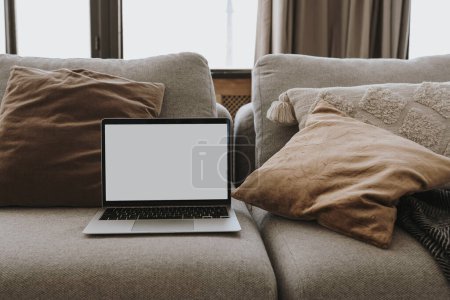 Laptop-Computer mit leerem Kopierplatz-Bildschirm auf einem bequemen Sofa mit Kissen. Ästhetische Wohnzimmereinrichtung. Online-Shopping, Online-Shop, soziale Medien, Blog-Branding-Attrappe