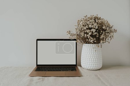 Foto de Ordenador portátil con pantalla en blanco en la mesa con ramo de flores de manzanilla. Plantilla de diseño de interiores de espacio de trabajo estilo influencer estético con espacio de copia de maqueta - Imagen libre de derechos
