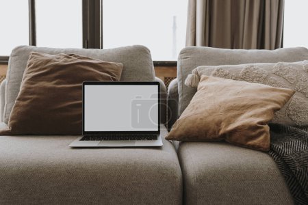 Laptop-Computer mit leerem Bildschirm auf Sofa mit Kissen. Ästhetisch elegant gestylte Wohnzimmereinrichtungsvorlage mit Kopiervorlage. Online-Shop, Blog, Social Media, Shop Branding