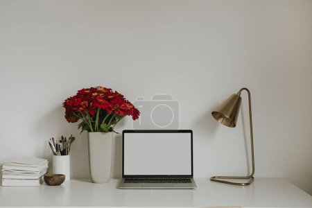 Foto de Ordenador portátil con pantalla en blanco en la mesa con flores de gerber rojo ramo. Plantilla de diseño de interiores de espacio de trabajo estilo influencer estético con espacio de copia de maqueta - Imagen libre de derechos