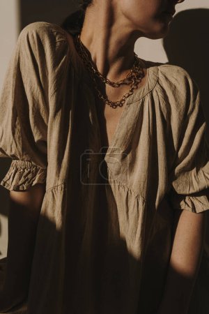 Téléchargez les photos : Jeune jolie femme en robe de lin et collier doré contre mur. Silhouette esthétique à la lumière du soleil. Ombres de lumière du soleil, nuances sur le mur. Concept élégant de conception de mode minimale - en image libre de droit