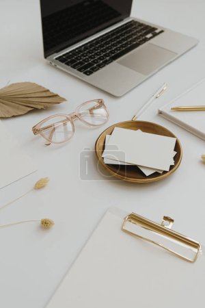 Photo for Home office desk workspace with laptop computer, blank paper card sheet with copyspace, glasses, stationery on white table. Women's, lady boss work, business background - Royalty Free Image