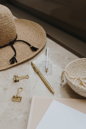 Gold female accessories and office stuff. Comfortable home office workspace. Work at home. Straw hat, clips, pen on marble table