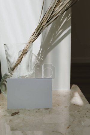 Téléchargez les photos : Feuille de carte papier vierge avec espace de copie vide. Feuille de palmier séchée dans un vase élégant sur une table en marbre. Des ombres sur le mur. Silhouette au soleil. Carte d'invitation de marque avec espace de copie - en image libre de droit