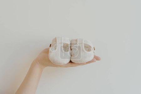 Téléchargez les photos : Tenez à la main une paire de petites sandales pour bébé mignonnes sur fond blanc. Composition minimale de la mode de bébé neutre pour le genre - en image libre de droit