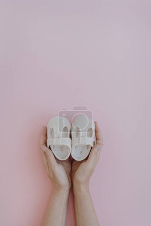 Photo for White cotton cute baby shoes in women's hands on pastel pink background. Fashion baby clothes - Royalty Free Image