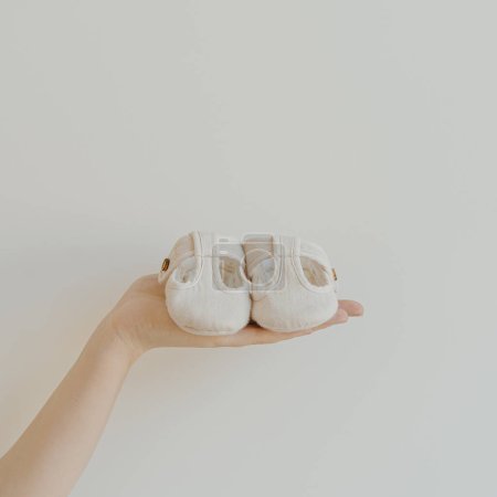 Téléchargez les photos : White cotton cute baby shoes in women's hand on white background. Fashion gender neutral baby clothes - en image libre de droit