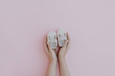 Téléchargez les photos : Hands hold pair of small cute newborn baby sandals shoes on pink background. Minimalist baby fashion composition - en image libre de droit