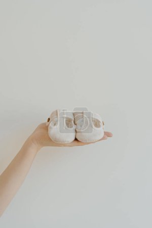 Photo for Person's hand hold cute little baby sandals shoes on white background. Minimalist baby fashion apparel - Royalty Free Image