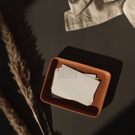 Photo for Flatlay of blank paper cards, plate, linen cloth, pampas grass, sunlight shadows on dark background. Business template. Top view, flat lay aesthetic luxury bohemian business branding concept - Royalty Free Image