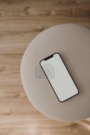 Téléchargez les photos : Téléphone portable à écran blanc avec espace de copie sur tabouret rond beige. Couché à plat, vue de dessus. Modèle de maquette d'espace de copie. - en image libre de droit