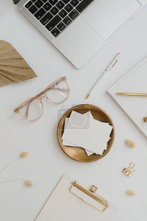 Photo for Home office desk workspace with laptop computer, blank paper card sheet with copyspace, glasses, stationery on white table. Flat lay, top view. Women's, girl boss work or business background - Royalty Free Image