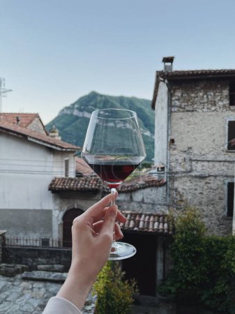 Foto de Mano de las mujeres sostienen la copa de vino con vino en frente de pintoresca vista tranquila de la antigua casa de campo italiana histórica en las montañas. Vacaciones de verano de lujo chill, relax - Imagen libre de derechos