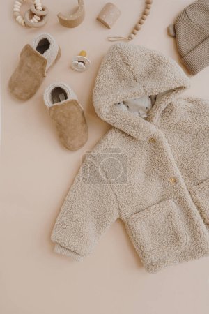 Photo for Stylish elegant baby clothes, accessories and toys on neutral beige background. Warm winter fleece jacket, suede booties, hat, pacifier, toys. Neutral pastel beige colour. Flat lay, top view - Royalty Free Image