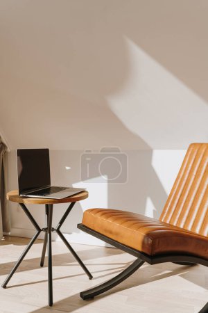 Téléchargez les photos : Esthétique luxe moderne espace de travail de bureau à domicile intérieur avec ordinateur portable, fauteuil en cuir orange et table basse. Ombres ensoleillées sur le mur. Espace lounge pour se détendre et se détendre - en image libre de droit