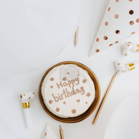 Téléchargez les photos : Gâteau d'anniversaire avec signe "Joyeux Anniversaire", ballons, bougies, chapeaux de fête. Couleurs blanc et or. Couché plat, vue du dessus. - en image libre de droit