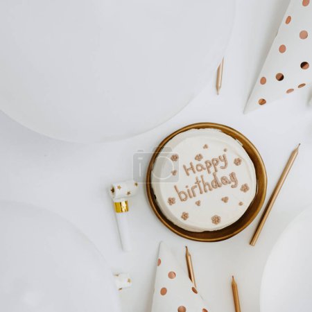 Photo for Elegant birthday cake with sign "Happy Birthday", candles, balloons, festive cones on white background. Aesthetic holiday event celebration concept. Flat lay, top view. White and gold colours - Royalty Free Image