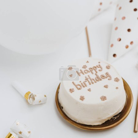 Foto de Elegante pastel de cumpleaños con signo "Feliz cumpleaños", velas, globos, conos festivos sobre fondo blanco. Concepto de celebración de eventos festivos estéticos. Colores blanco y dorado - Imagen libre de derechos