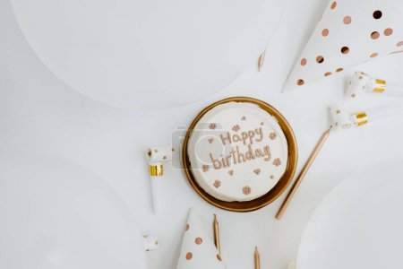 Téléchargez les photos : Gâteau d'anniversaire avec signe "Joyeux Anniversaire", ballons, bougies, chapeaux de fête. Couleurs blanc et or. Couché plat, vue du dessus. - en image libre de droit