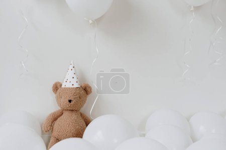 Téléchargez les photos : Jolie peluche ours en chapeau de fête assis près de ballons sur fond blanc. Fête d'anniversaire des enfants. Le premier anniversaire du bébé. Ballon avec espace de copie vierge - en image libre de droit