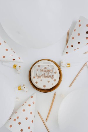 Foto de Tarta de cumpleaños, velas, globos, sombreros de fiesta sobre fondo blanco. Asiento plano, vista superior. Colores blanco y dorado - Imagen libre de derechos