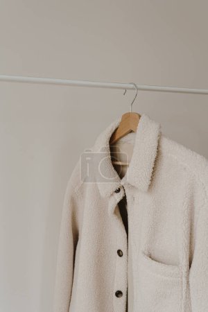 Foto de Ropa de abrigo de otoño en percha sobre pared blanca. Chaqueta blanca cálida. Armario estético con ropa en colores neutros - Imagen libre de derechos