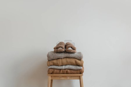 Téléchargez les photos : Empilement de vêtements beiges neutres chauds sur tabouret en bois au-dessus du mur blanc. Babouches moelleuses, pulls et pulls en laine - en image libre de droit