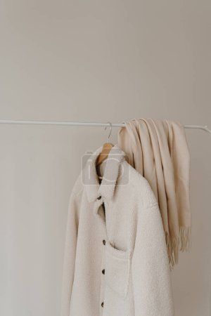 Foto de Ropa de abrigo de otoño en percha sobre pared blanca. Chaqueta blanca cálida y falda. Armario estético con ropa en colores neutros - Imagen libre de derechos