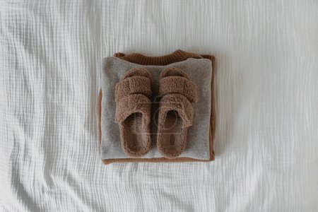 Téléchargez les photos : Des pantoufles brunes confortables sur une pile de pulls de laine chaude et des pulls sur un tissu de mousseline blanche. Porté chaud à la maison. Couché plat, vue du dessus - en image libre de droit