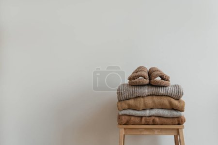 Photo for Stack of warm neutral beige clothes on wooden stool over white wall. Fluffy slippers, wool pullovers and sweaters. Minimalist autumn, fall fashion background - Royalty Free Image