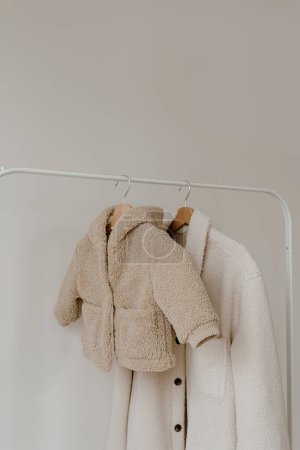 Foto de Cálido otoño bebé de temporada y ropa de adulto en percha sobre la pared blanca. Chaquetas de lana neutra beige, blanca y negra. Armario de ropa de moda minimalista - Imagen libre de derechos