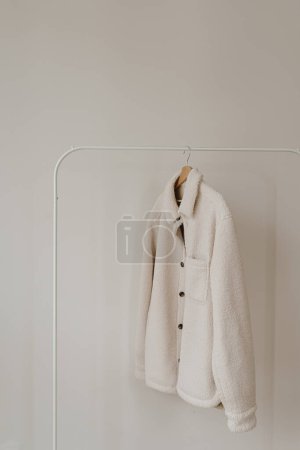 Téléchargez les photos : Veste saison automne en laine chaude blanche sur cintre au-dessus du mur blanc. Armoire minimaliste de vêtements de mode - en image libre de droit