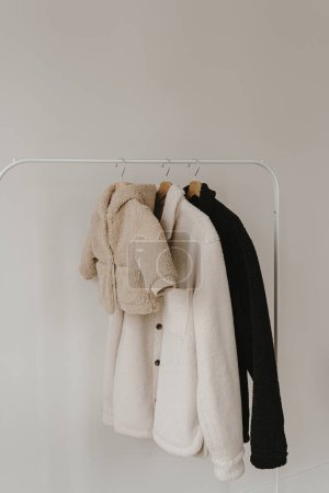 Foto de Cálido otoño bebé de temporada y ropa de adulto en percha sobre la pared blanca. Chaquetas de lana neutra beige, blanca y negra. Armario de ropa de moda minimalista - Imagen libre de derechos