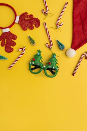 Téléchargez les photos : Nouvel An drôle, accessoires de Noël pour la fête des vacances d'hiver fête sur fond jaune. Lunettes de Noël, cannes à bonbons, cornes de cerf, chapeau de Père Noël, arbres jouets - en image libre de droit