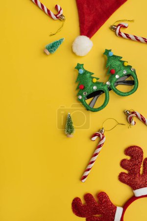 Téléchargez les photos : Lunettes de Noël drôles colorées, cannes à bonbons, cornes de cerf, chapeau de Père Noël, sapins jouets sur fond jaune. Nouvel An, Accessoires de Noël pour la fête des vacances d'hiver - en image libre de droit