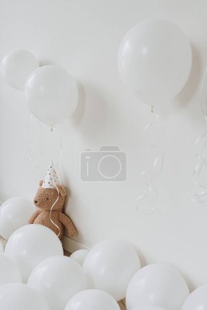Foto de Osito de peluche en sombrero de fiesta sentado cerca de globos sobre fondo blanco. Fiesta de cumpleaños para niños. Primer aniversario del bebé - Imagen libre de derechos