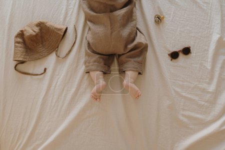 Photo for Newborn baby in stylish basic bodysuit with accessories on neutral beige bed linens. Fashion Scandinavian newborn clothes. Hat, pacifier, sunglasses. Flat lay, top view - Royalty Free Image