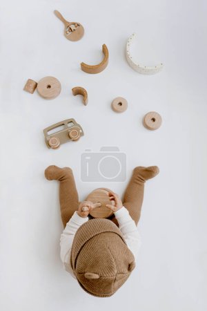 Téléchargez les photos : Petit bébé dans des vêtements élégants joue avec des jouets en bois. Mode Scandinave vêtements et jouets pour nouveau-nés. Couché plat, vue du dessus - en image libre de droit