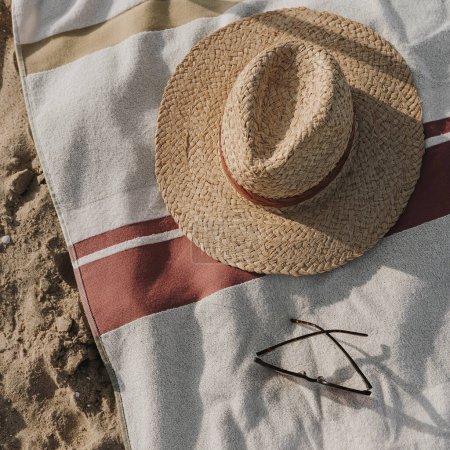 Téléchargez les photos : Chapeau de paille, lunettes de soleil, serviette de plage sur sable de plage. Mode féminine minimaliste fond de vogue. Couché plat, vue du dessus - en image libre de droit