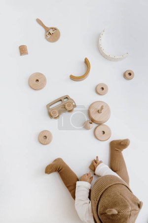 Photo for Little baby in stylish clothes plays with wooden toys. Fashion Scandinavian newborn clothes and toys. Hat, jumpsuit, bodysuit. Flat lay, top view - Royalty Free Image